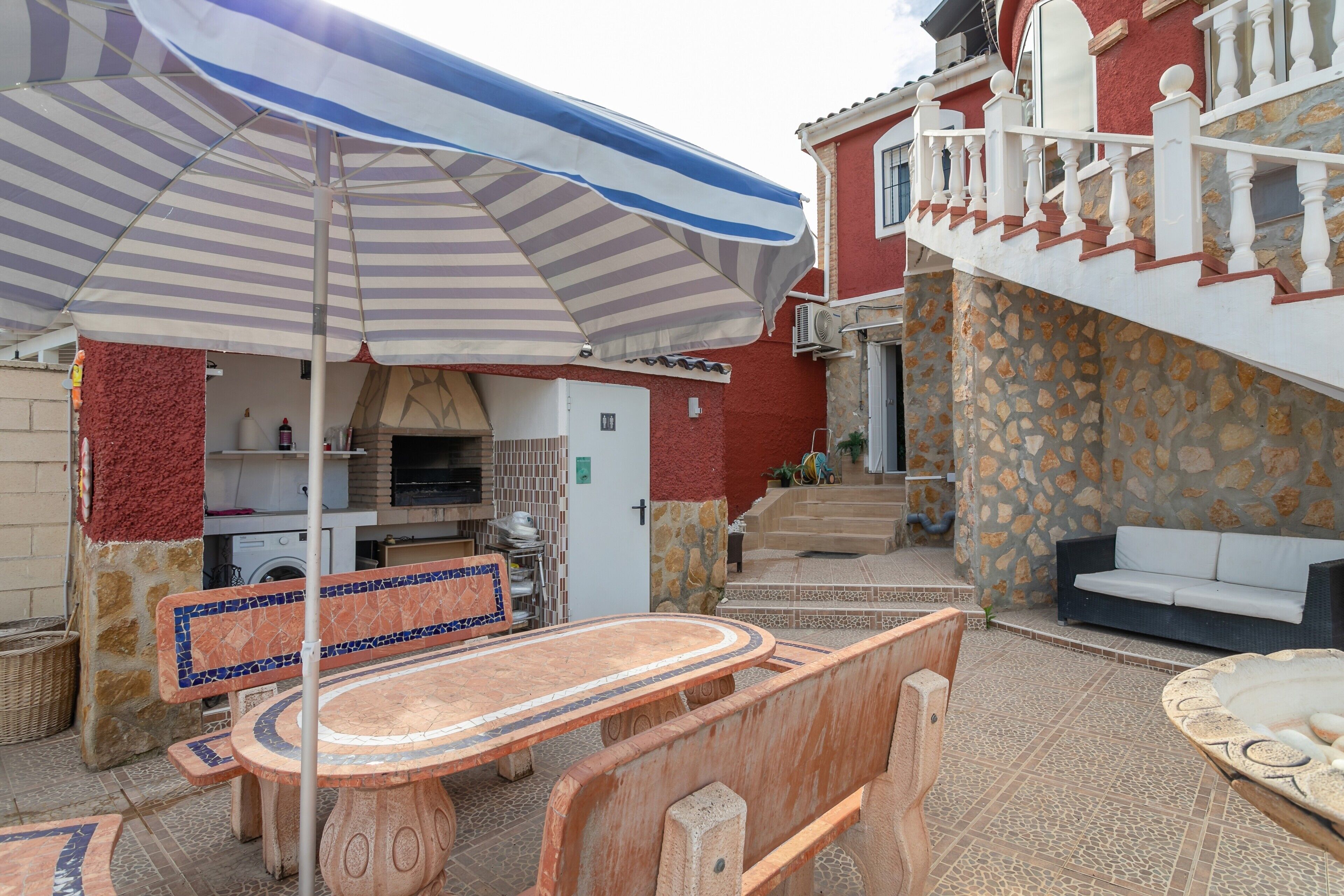 A patio with a table and chairs, a striped umbrella, a stone wall, and a staircase leading to a balcony.