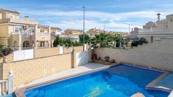 Una piscina con un borde decorativo de azulejos, rodeada de muros de ladrillo color crema y una valla blanca.