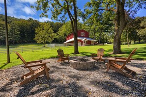A rustic outdoor seating area with Adirondack chairs and a fire pit, surrounded by a white fence and a red barn in the background. - Lazy 8 is a remodeled historical farmhouse in the countryside of Snyder Co., PA (Beavertown)