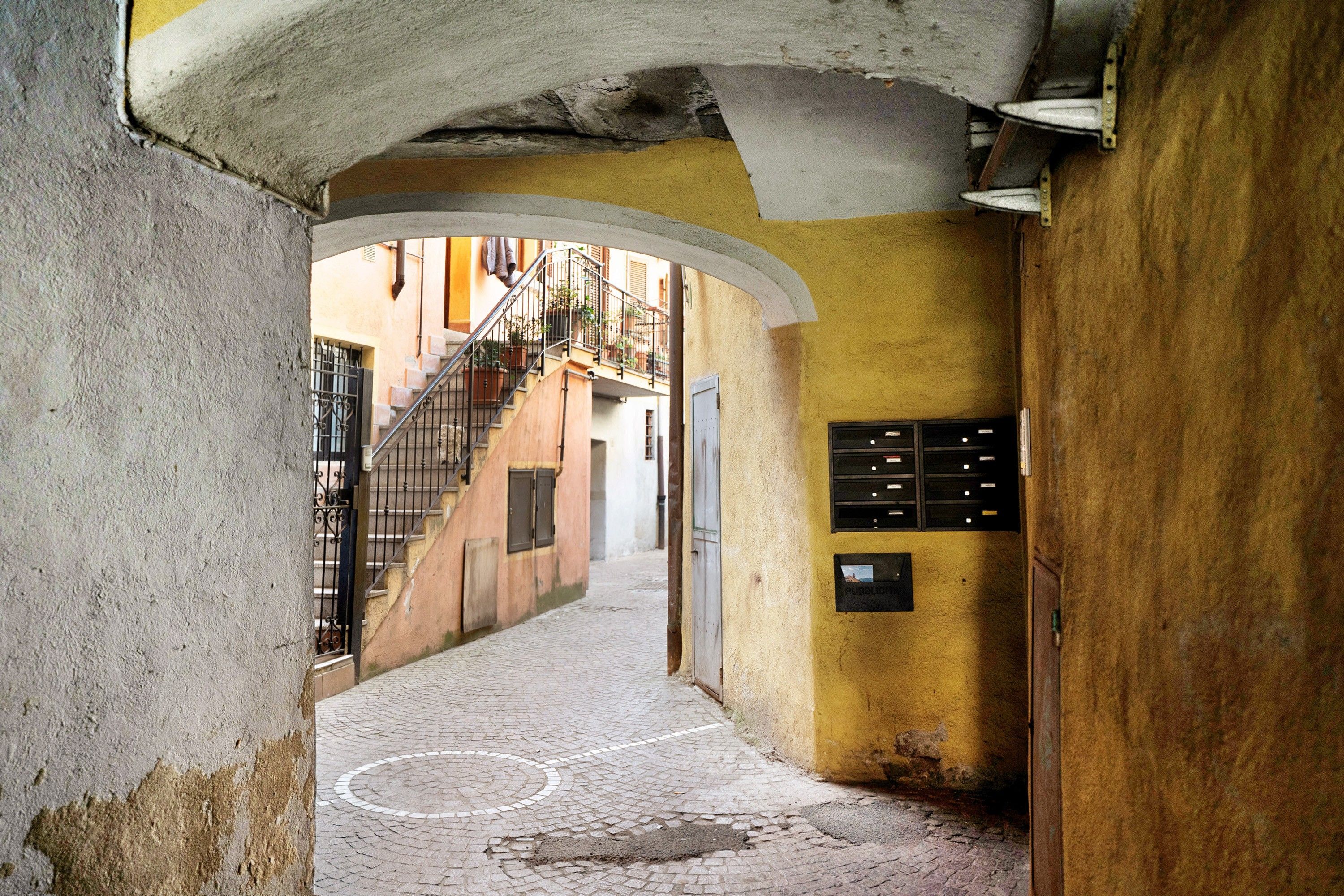 A narrow alleyway with a cobblestone path, a metal gate, and a staircase leading to a building with a yellow wall and a mailbox.
