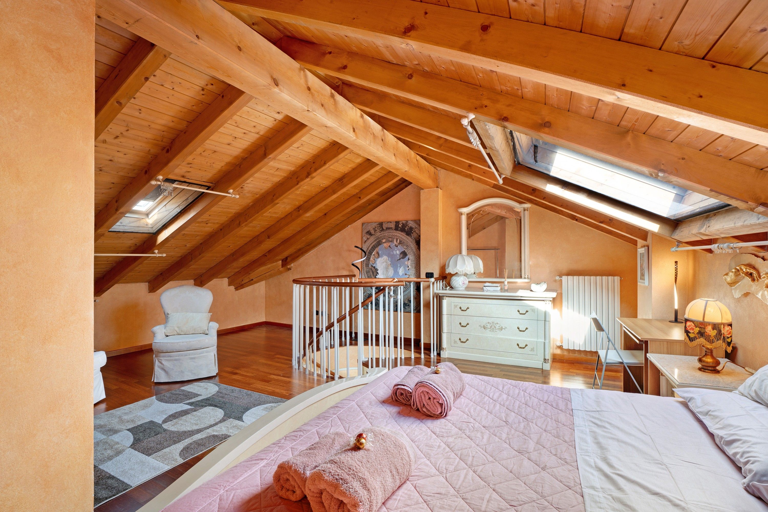 A bedroom with a sloped ceiling, wooden beams, a bed with a quilt, a chair, a dresser, and a mirror.