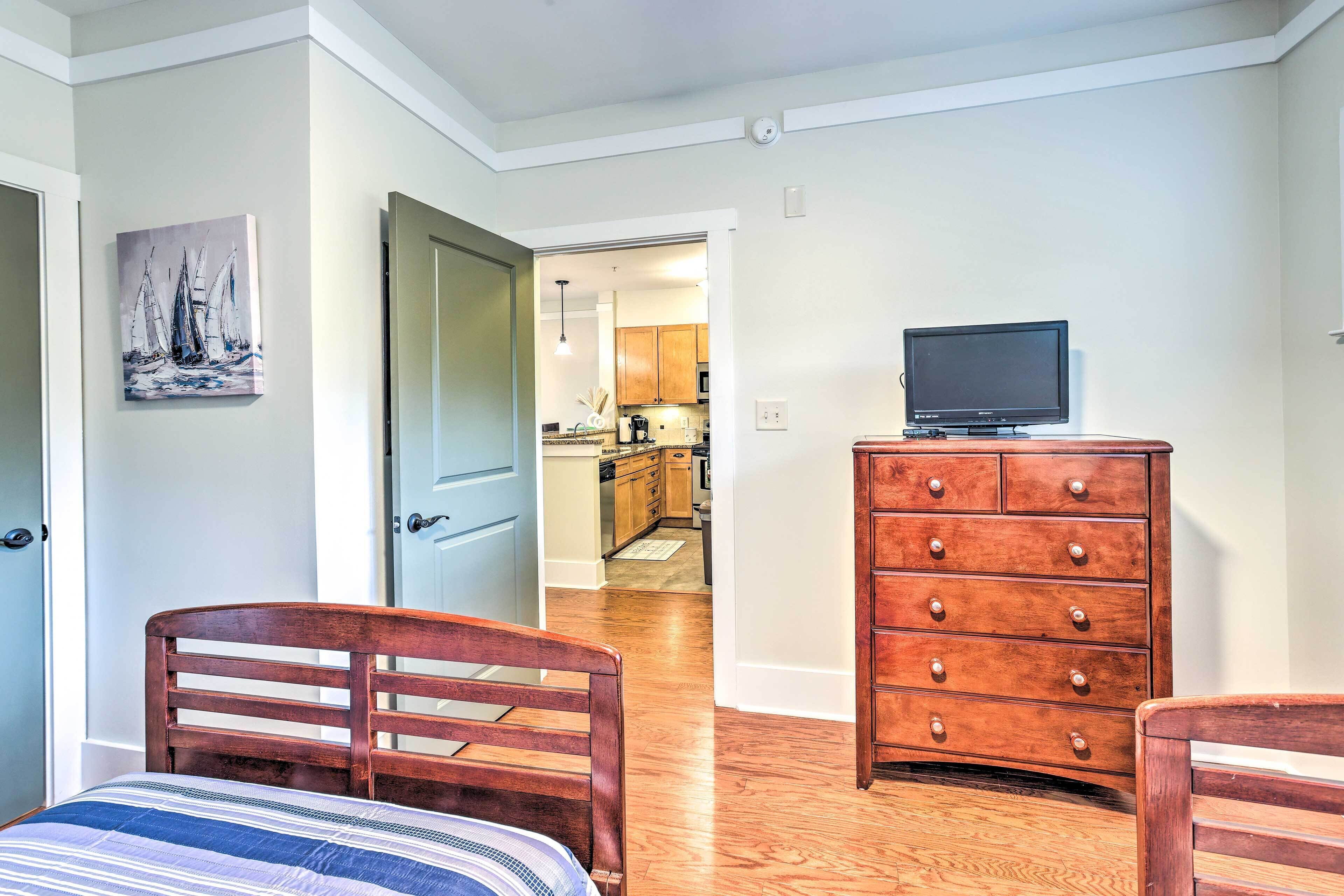 Une chambre avec un lit en bois, une commode et un téléviseur posé sur une autre commode. Il y a un tableau accroché au mur et une porte qui donne sur la cuisine.