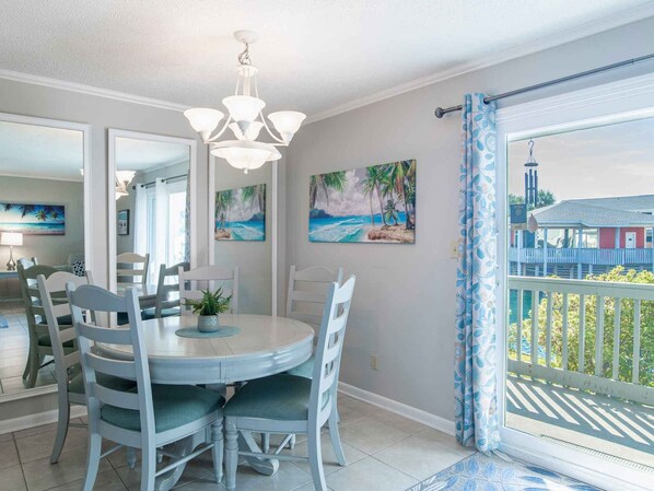 Un coin repas avec une table ronde, des chaises blanches et un lustre. Deux tableaux représentant des scènes tropicales ornent le mur.