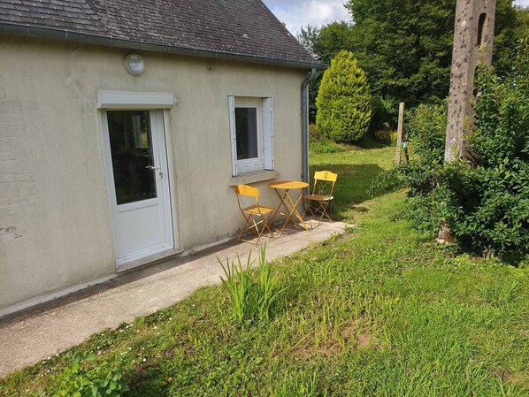 Un bâtiment d’un seul étage, doté d’une porte et de deux fenêtres, un petit espace extérieur aménagé avec deux chaises jaunes, et une cour verdoyante avec des arbres et des arbustes.