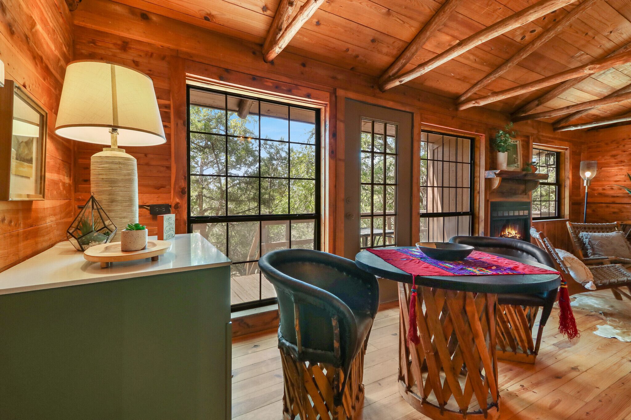 A wooden cabin interior with a dining table, chairs, a lamp, and a fireplace.