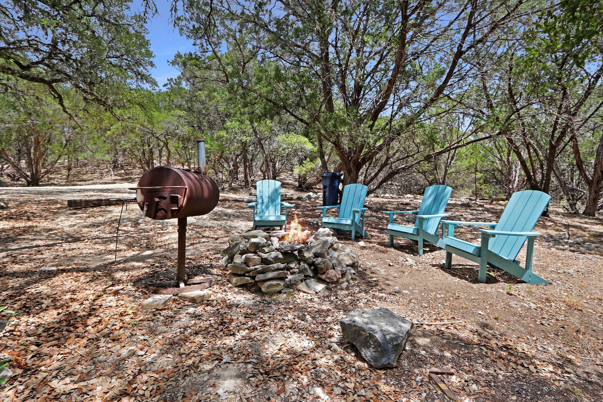 A campsite with a fire pit, a metal barrel, and several blue Adirondack chairs.