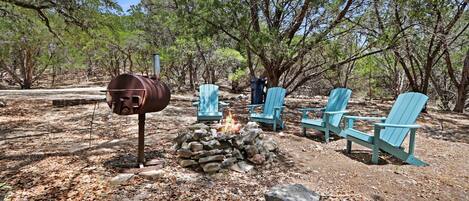 A campsite with a fire pit, a metal barrel, and several blue Adirondack chairs.