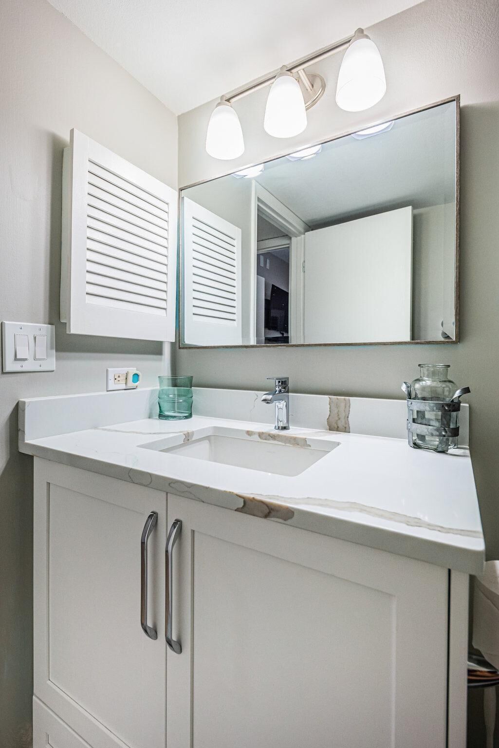 Une salle de bain avec un meuble-lavabo blanc, un évier, un miroir et un luminaire placé au-dessus du miroir.