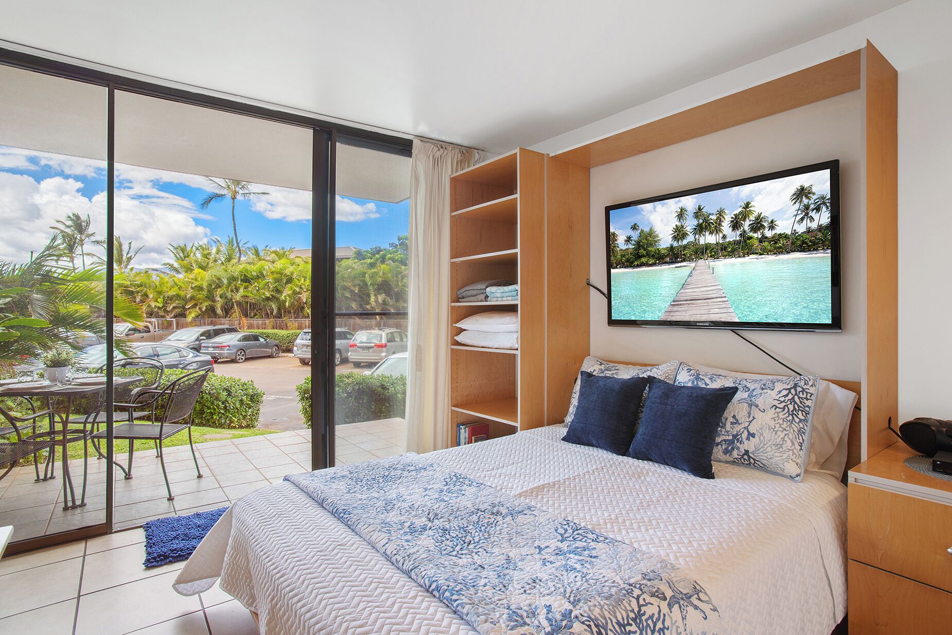A bedroom with a bed, a TV, a sliding glass door to a balcony, and a view of a beach.