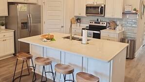 Une cuisine avec des armoires blanches, un réfrigérateur en acier inoxydable, un four à micro-ondes et une cuisinière. On y trouve des tabourets en bois près de l’îlot central.