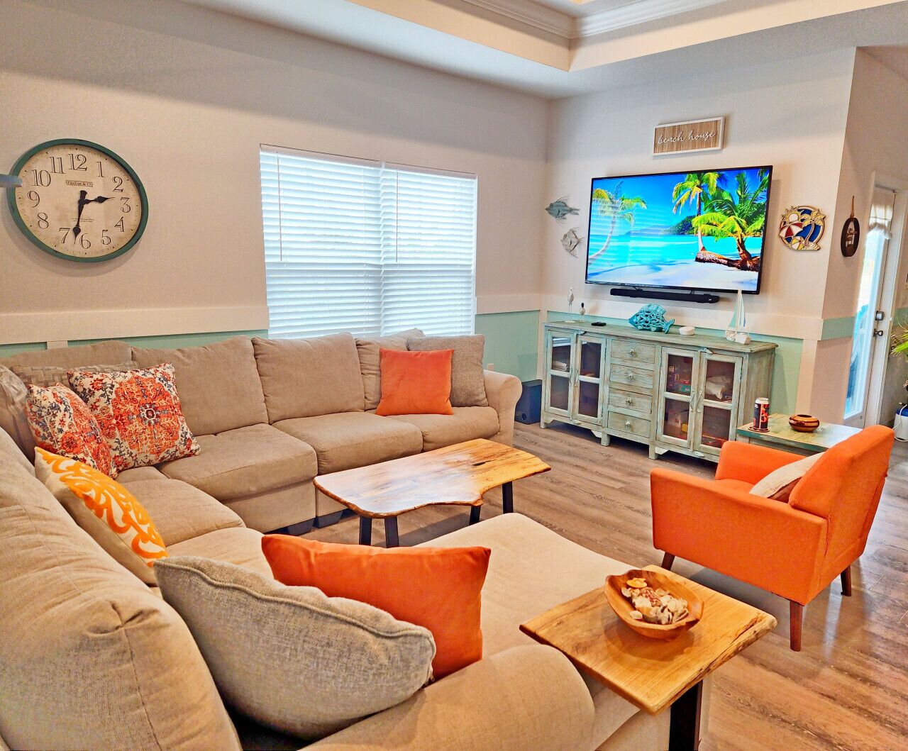 Un salon avec un canapé beige, des fauteuils orange, une table basse en bois et un téléviseur diffusant une scène tropicale.