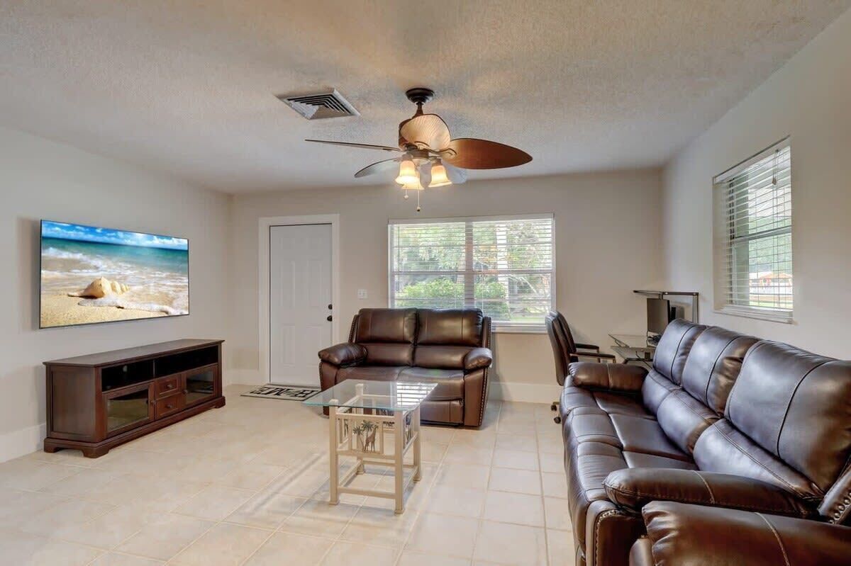 A living room with a ceiling fan, a brown leather sofa, a glass coffee table, a brown leather armchair, a TV on the wall, and a window with blinds.