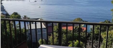 A balcony with a table and chairs, overlooking a calm sea with boats and distant land.