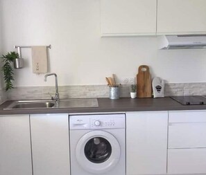 A compact kitchen with a built-in washing machine, a stainless steel sink, and a wooden cutting board. - Elegance - Sea view, private beach access, swimming pool (Schoelcher)