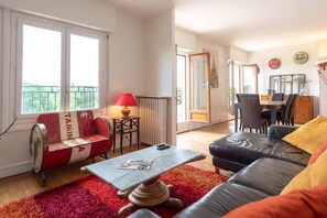 Un salon avec un banc rouge et blanc, un canapé en cuir noir, un tapis rouge et un coin repas comprenant une table et des chaises.