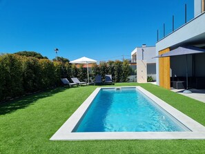Una piscina rectangular con borde blanco, rodeada de césped artificial verde. Se ven dos sillas de descanso y una sombrilla blanca al fondo.