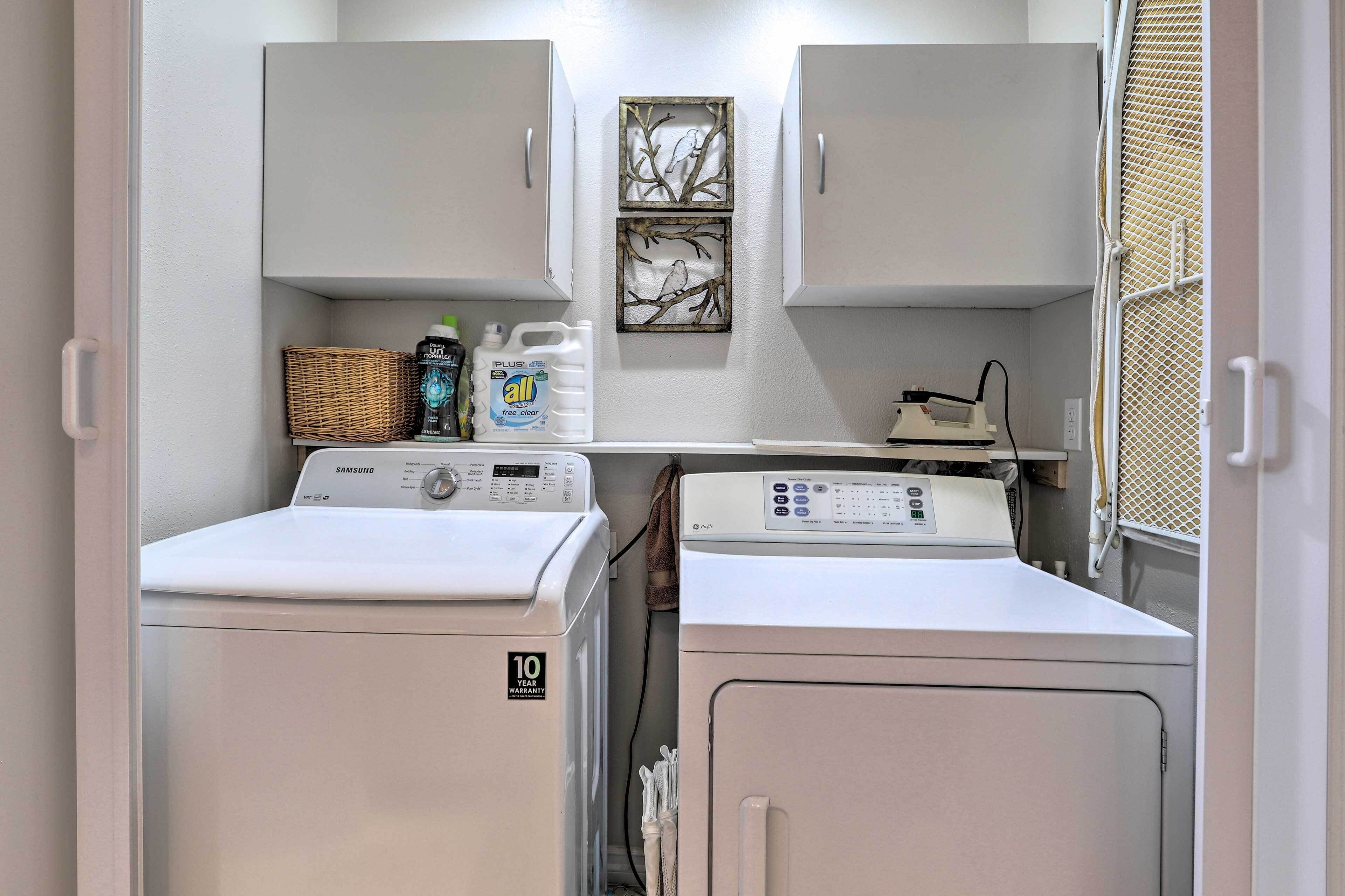 A laundry room with two washing machines, a dryer, and a shelf with cleaning supplies.