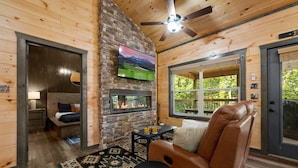 A cozy cabin interior with a stone fireplace, a ceiling fan, a large window, and a leather recliner. - Private Location Hot tub, Pool table, Multicade, Free Attraction Passes (Sevierville)