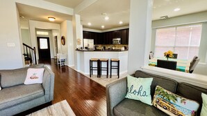 A spacious living room with a grey sofa and a grey couch, both with decorative pillows. A kitchen area with dark cabinets and a central island is visible in the background.