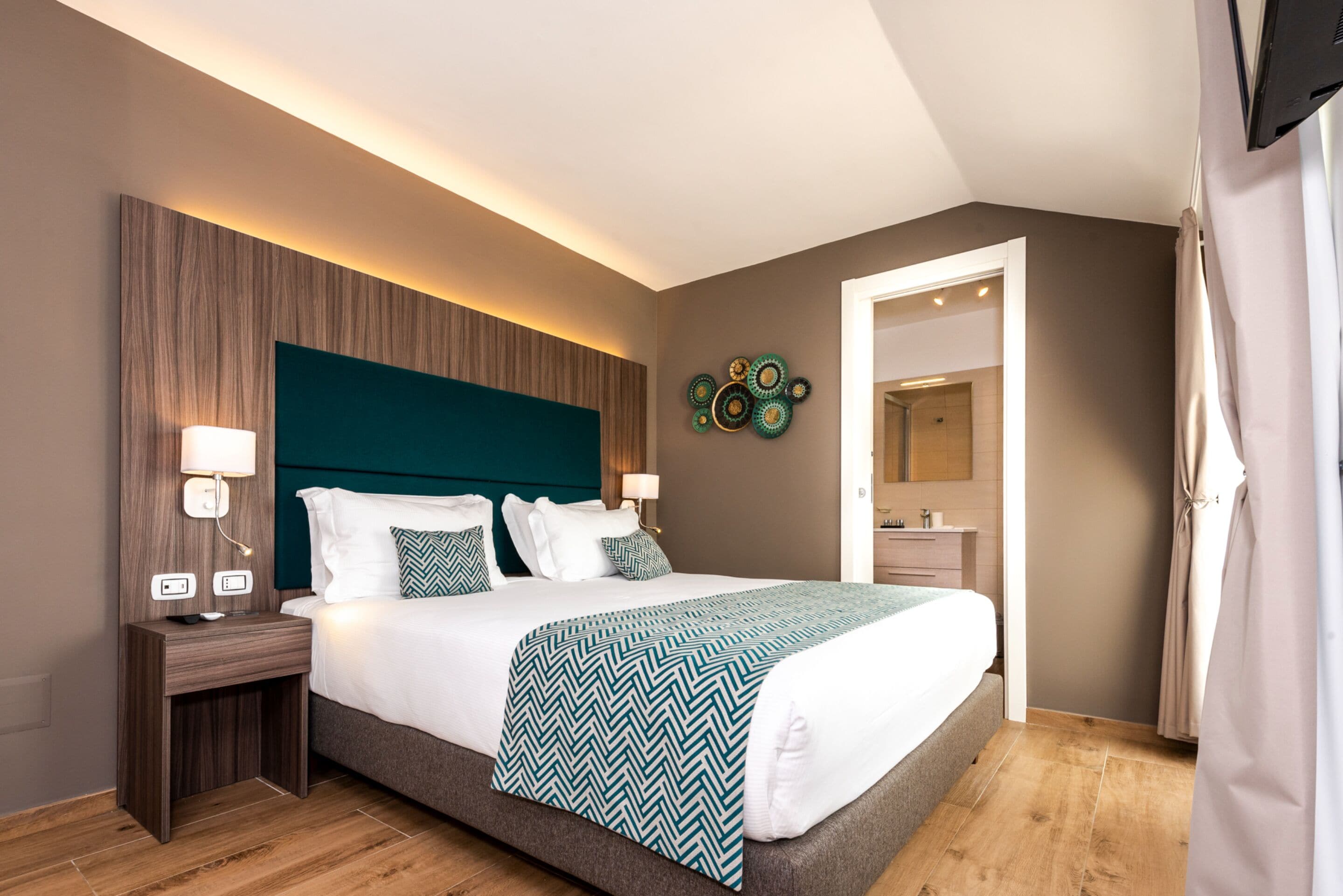 A modern hotel room with a large bed, bedside tables with lamps, a headboard, and a decorative wall piece.