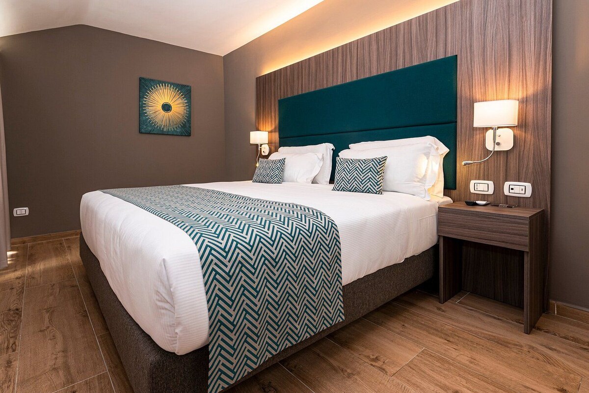Hotelkamer met een groot bed, twee nachtkastjes, een wandlamp en een decoratief object aan de muur.