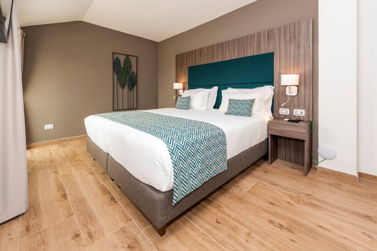Een moderne hotelkamer met een groot bed, houten vloer, een blauwgroen hoofdeinde en bedlampjes.