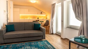 A modern living room with a grey sofa, teal cushions, a patterned rug, a dining area with chairs, and a kitchen with a countertop and appliances.