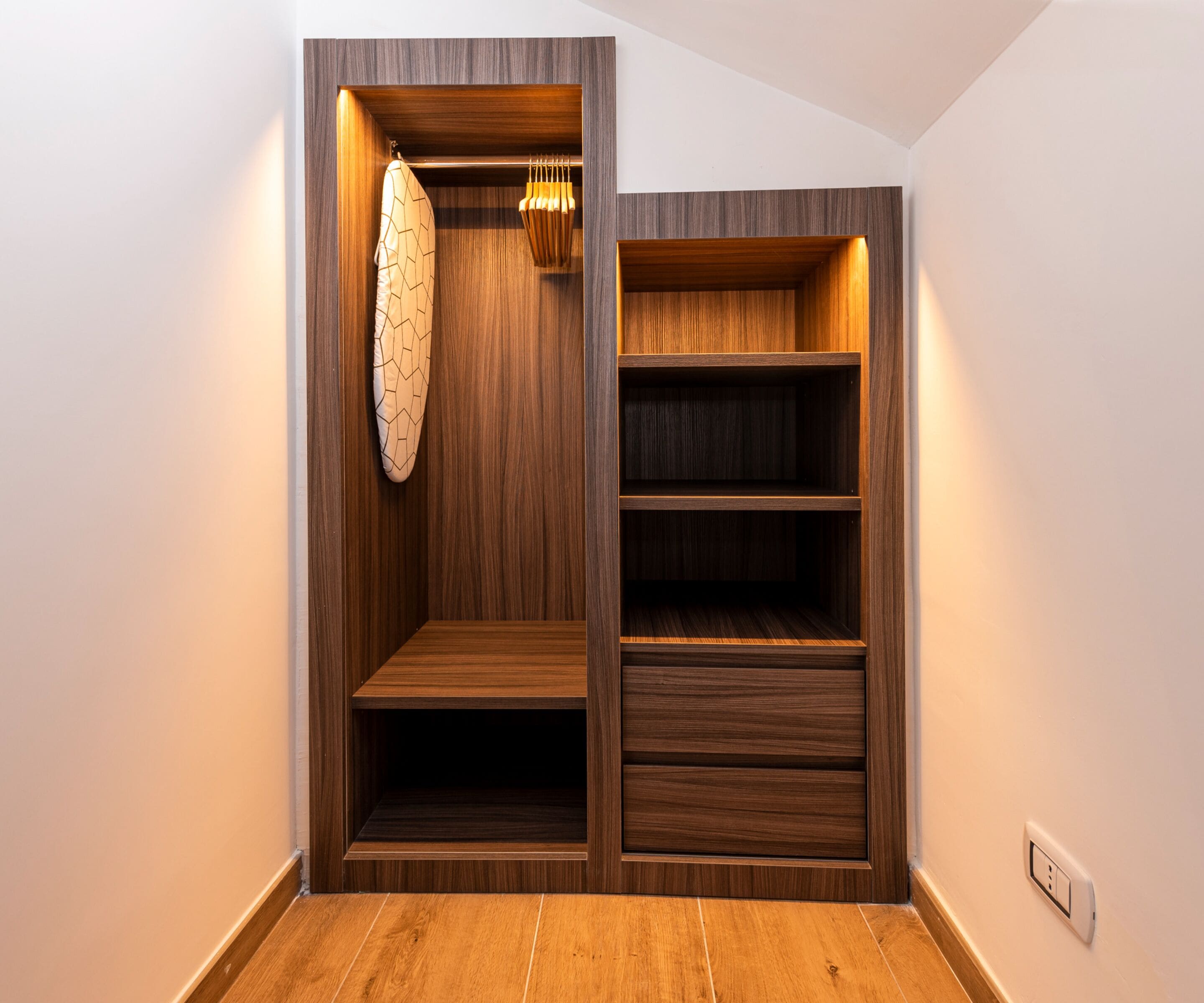 A wooden closet with shelves and hanging space, a light fixture, and a power outlet on the wall.