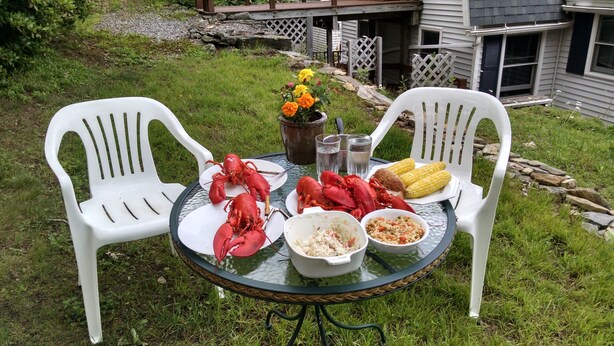A round glass table with white plastic chairs, a plate of lobsters, corn on the cob, and bowls of food. - Classic Maine House, wrap around deck,  water activity a walk away. (Harpswell)