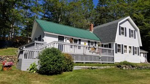 A two-story house with a green roof, a porch with a railing, and a stone wall. - Classic Maine House, wrap around deck,  water activity a walk away. (Harpswell)