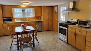 A kitchen with wooden cabinets, a microwave, a stove, a toaster, and a dining table with chairs. - Classic Maine House, wrap around deck,  water activity a walk away. (Harpswell)
