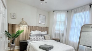 Une chambre avec un grand lit, un ventilateur de plafond, une table de chevet, une commode, une plante en pot et une fenêtre avec des rideaux.