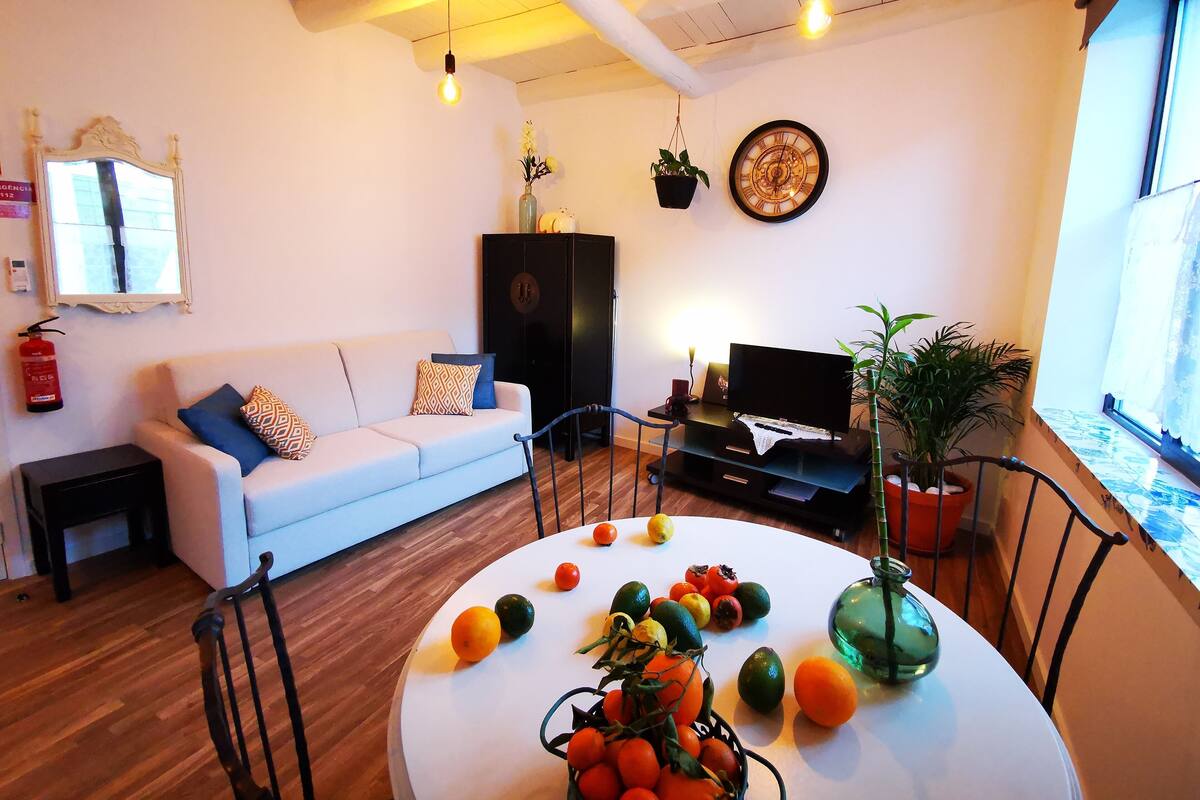 A living room with a white sofa, a black entertainment unit, a round table with fruit, and a large window.