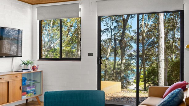 A modern living room with a large sliding glass door leading to a balcony with a scenic view of trees and a body of water. - Over The Boardwalk - North Narooma (North Narooma)