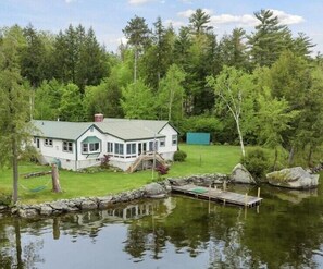 A house with a green roof, a porch, and a dock extending into a pond. - Lake Wentworth Estate: Unobstructed Sunset Views, Private Beach, Fire Pit, Lawn (Wolfeboro)