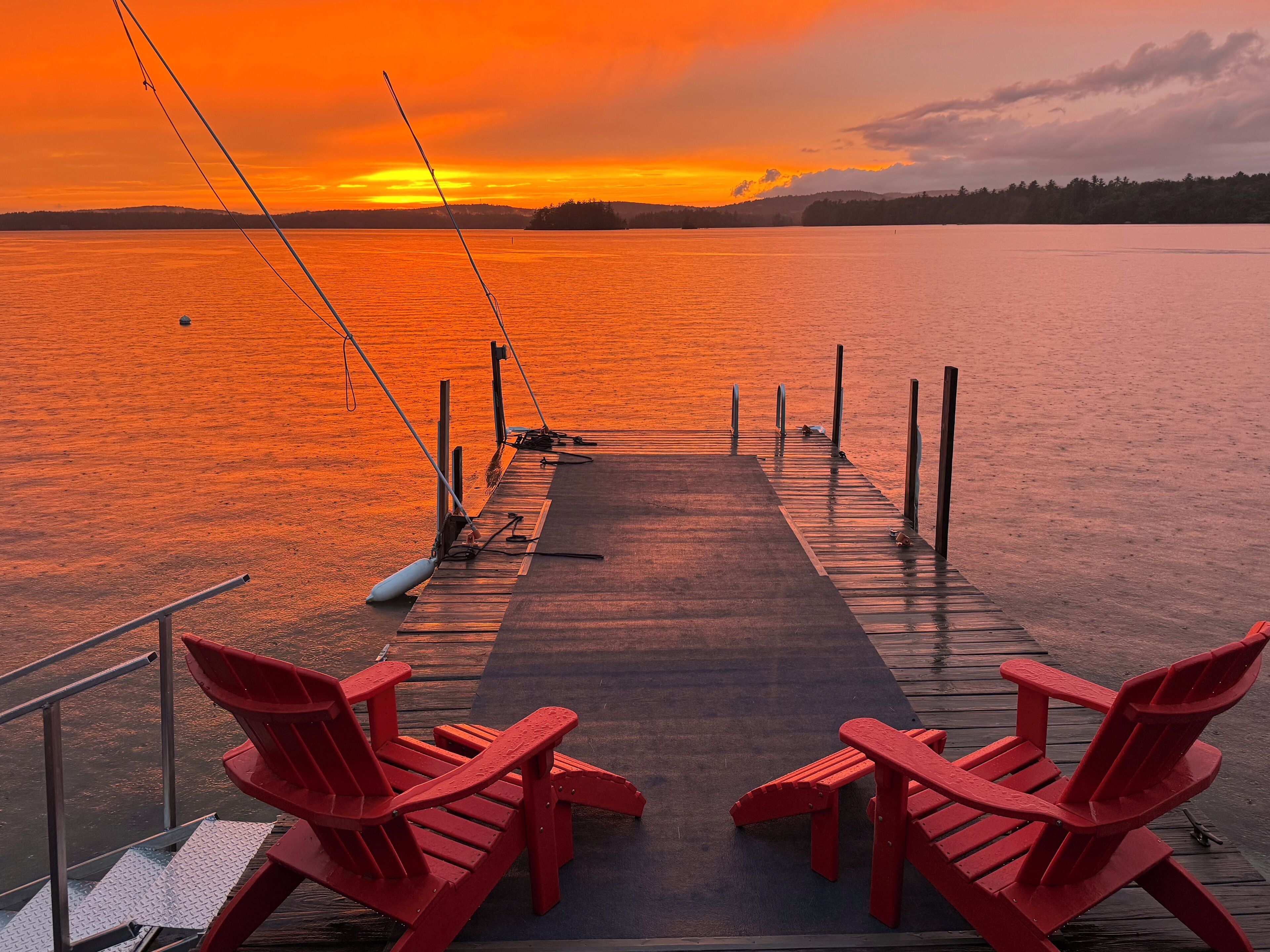 A wooden dock with two red Adirondack chairs facing a calm lake at sunset.