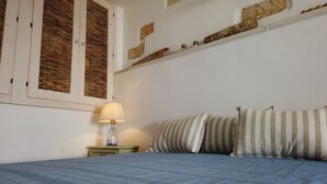A bedroom with a bed, striped pillows, a nightstand with a lamp, a window with bamboo blinds, and a shelf with decorative items. - Blue House (Castro)