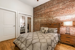Un dormitorio con pared de ladrillo, cama con ropa de cama estampada, mesita de noche de madera y puerta blanca.