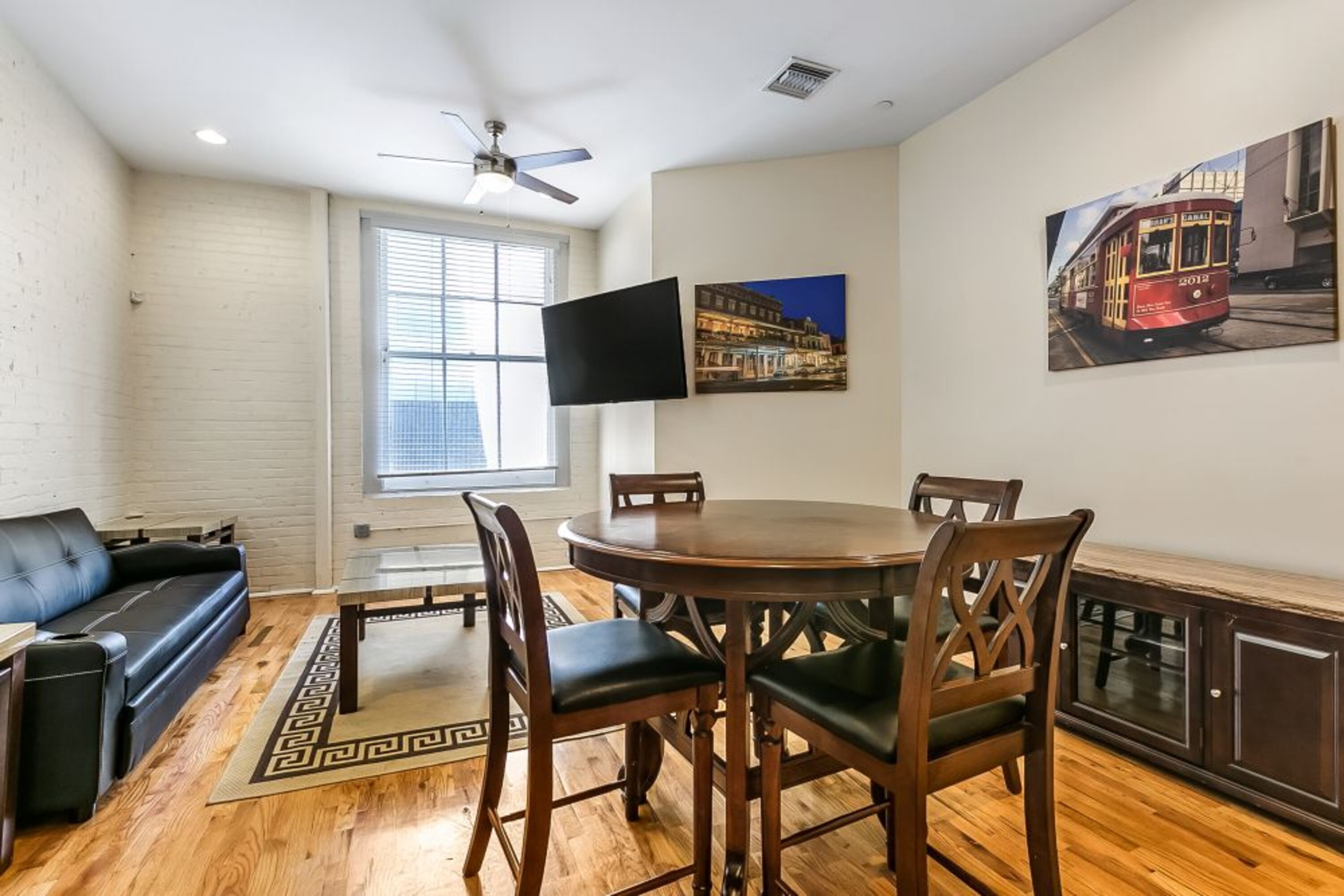 A dining area with a round table, chairs, a black sofa, a TV mounted on the wall, and a window with blinds.