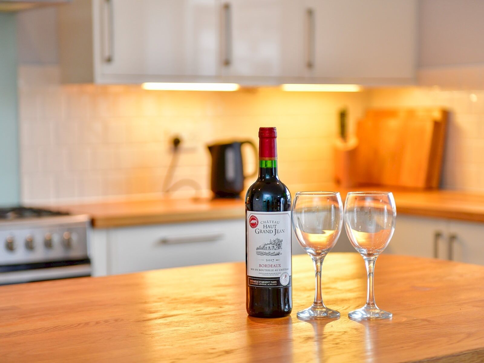 A bottle of Chateau Haut Grand Jean wine from 2017 on a kitchen counter with two empty wine glasses.