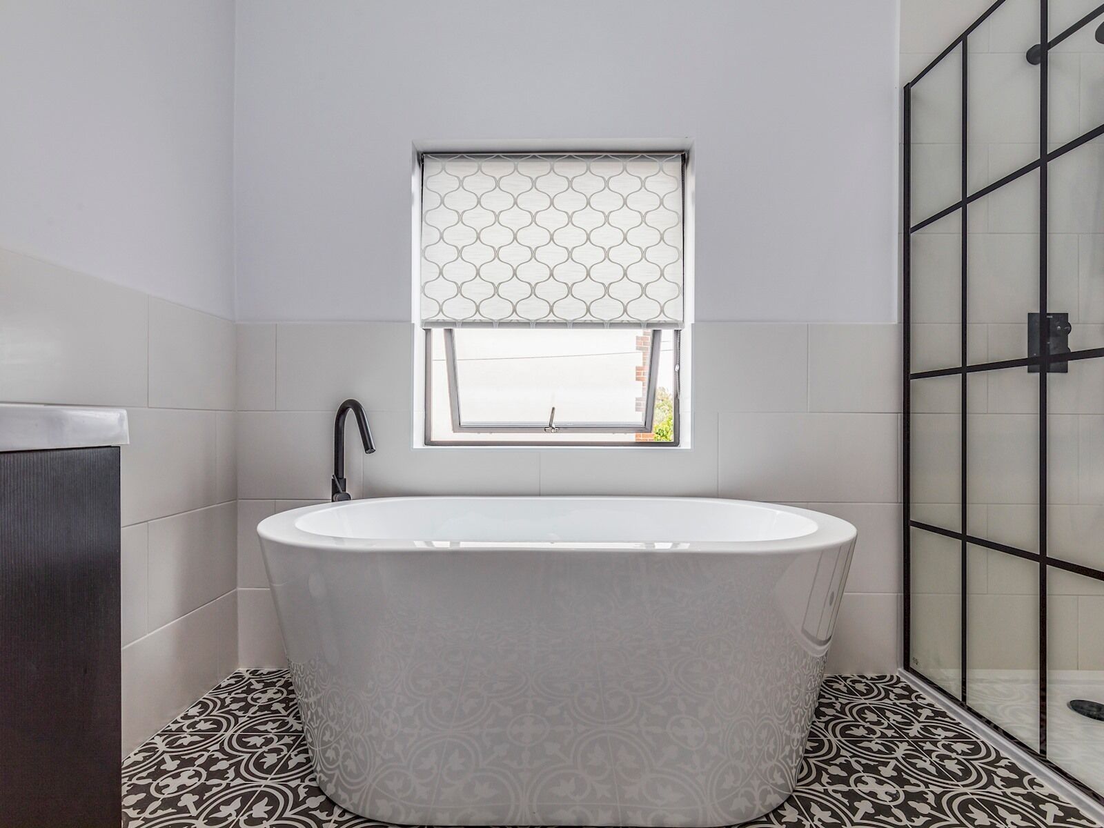 A modern bathroom with a freestanding bathtub, a window with a geometric patterned blind, and a shower area with a glass door.
