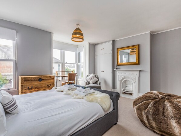 A bedroom with a bed, a wooden chest of drawers, a grey sofa, a brown pouf, a fireplace, and a large window.