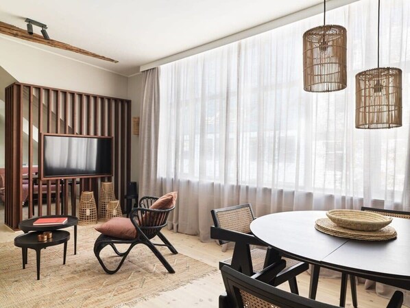 Un salon moderne avec une table à manger ronde, deux chaises, un téléviseur et une grande fenêtre agrémentée de rideaux légers.