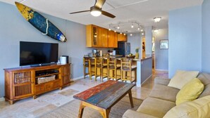 Una sala de estar con televisor, mueble de entretenimiento de madera, un banco, un sofá y una zona de cocina con armarios de madera y una barra.