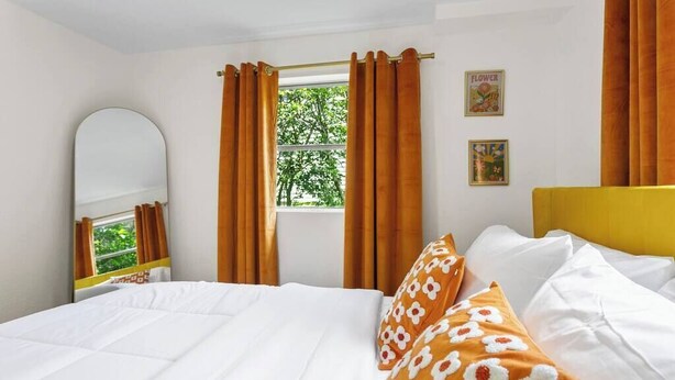 A bedroom with a bed, a window with curtains, a mirror, and a wall with two framed pictures. - That 70s Themed Apt Close to Coffee, Food and Shops (Tampa)