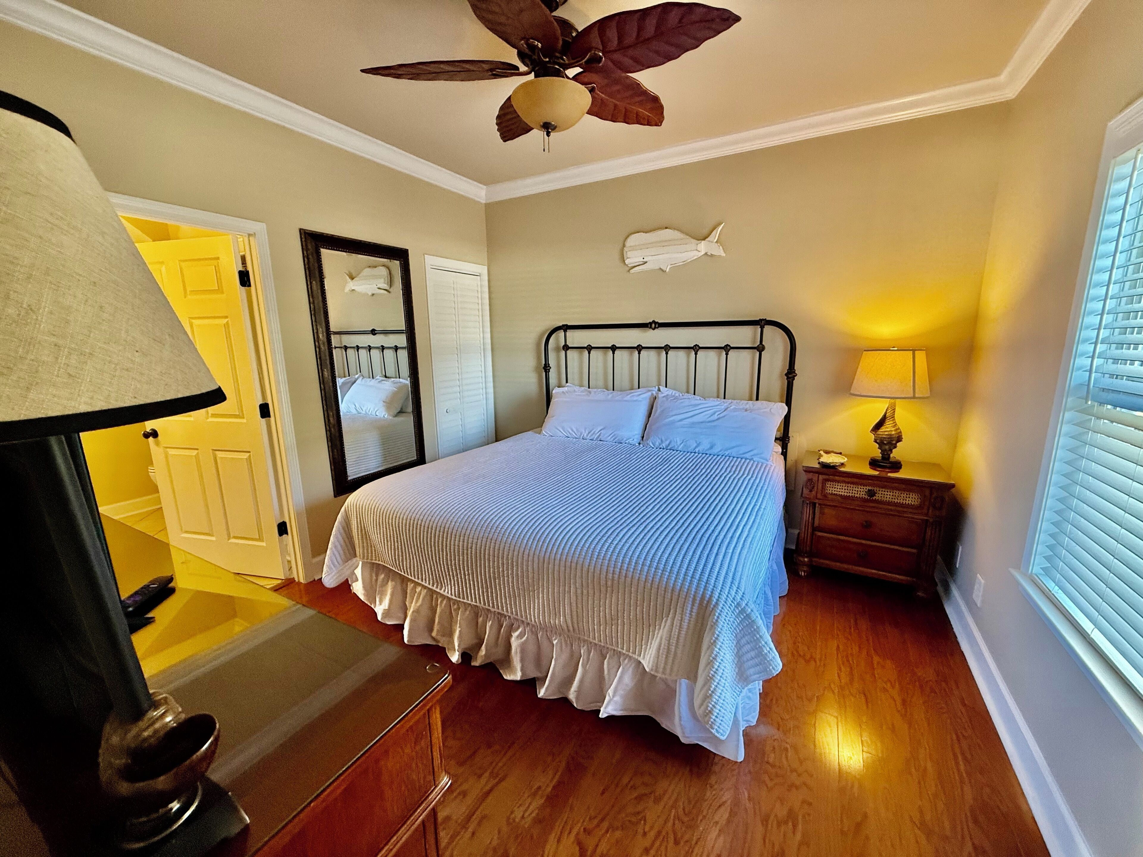 A bedroom with a bed, a ceiling fan, a nightstand, a lamp, a mirror, and a window with blinds.