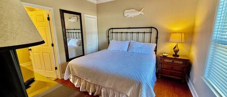 A bedroom with a bed, a ceiling fan, a nightstand, a lamp, a mirror, and a window with blinds.