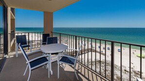 Un balcon avec une table ronde et des chaises, donnant sur une plage parsemée de parasols et fréquentée par des gens.