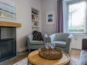 A living room with two armchairs, a fireplace, a bookshelf, and a window with curtains. - Parkview Cottage - Beautiful terraced bungalow with cosy fireplace (Carnoustie)