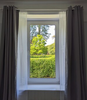 A window with grey curtains, a white frame, and a view of a lush garden with a large tree and neatly trimmed hedges. - Parkview Cottage - Beautiful terraced bungalow with cosy fireplace (Carnoustie)